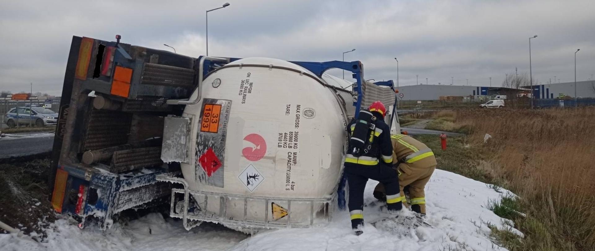 Biała cysterna leży przewrócona w rowie. Przy cysternie znajdują się dwaj strażacy. Jeden z nich ubrany w ubranie specjalne koloru czarnego, czerwony hełm. Na plecach ma założony aparat powietrzny. Drugo strażak pochylony przy cysternie, ubrany w ubranie specjalne w kolorze żółtym. Wokół cysterny na pasie zieleni i na powierzchni rowu widać podaną białą pianę gaśniczą.