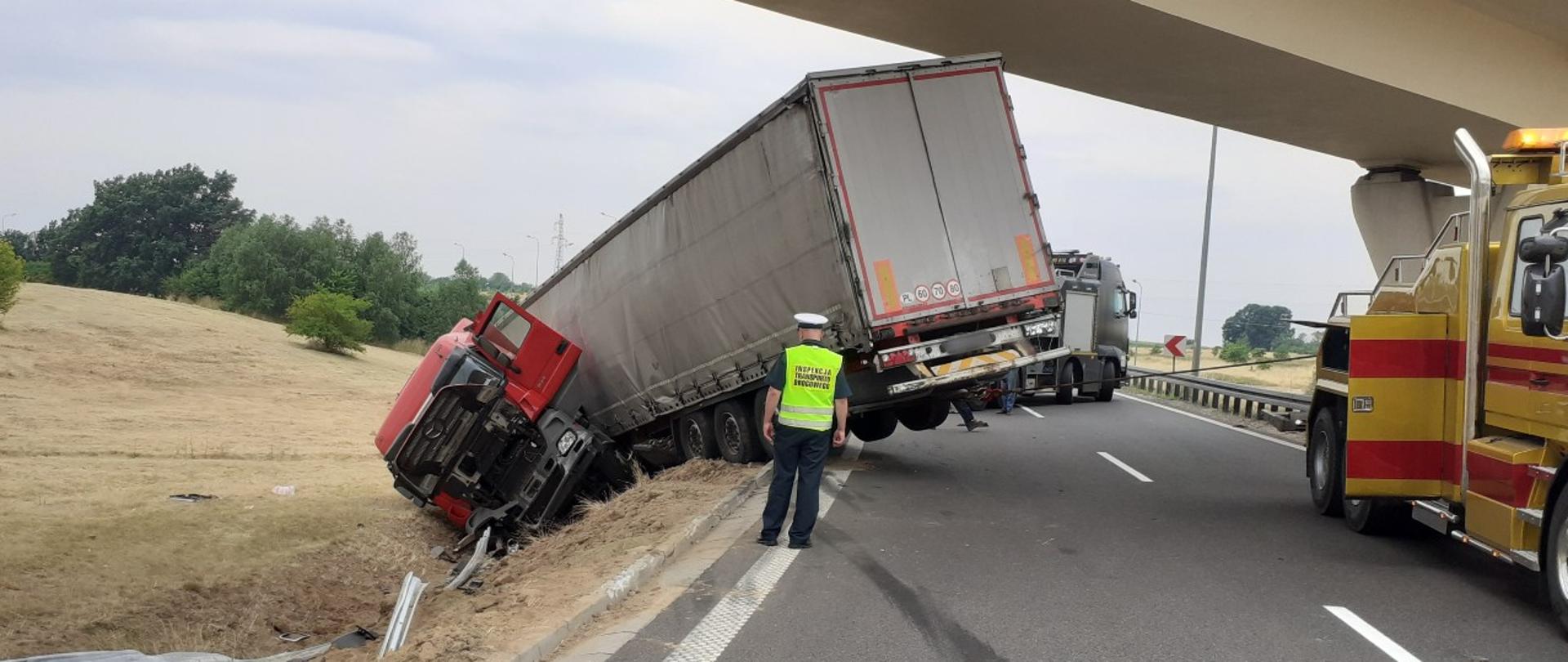 Droga z dwoma pasami ruchu, nad nią wiadukt. Na drodze stoi inspektor ITD i patrzy na ciężarówkę, która miała wypadek i leży częściowo na poboczu. Obok stoją dwa holowniki.