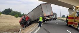 Droga z dwoma pasami ruchu, nad nią wiadukt. Na drodze stoi inspektor ITD i patrzy na ciężarówkę, która miała wypadek i leży częściowo na poboczu. Obok stoją dwa holowniki.