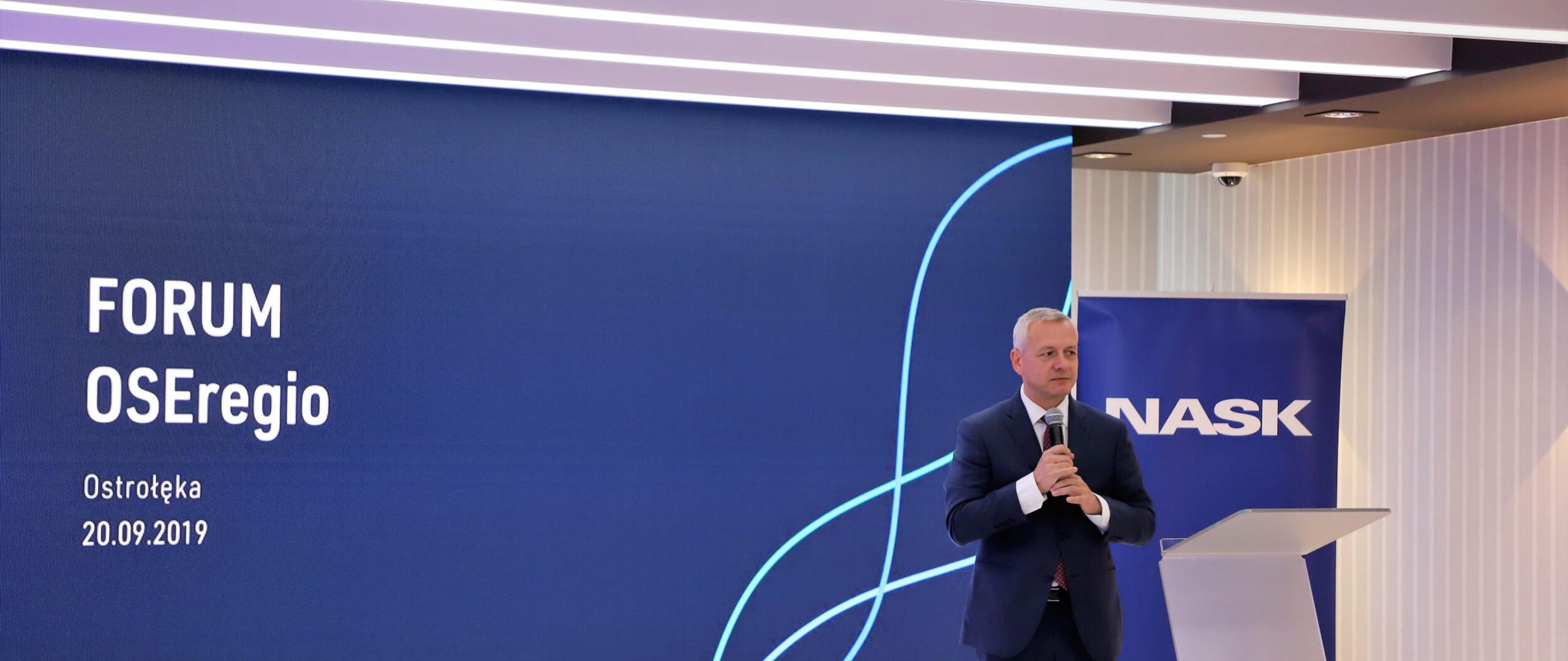 Minister Marek Zagórski trzyma mikrofon w rękach stojąc na tle ekranu, na którym wyświetla się nazwa i data wydarzenia oraz roll-upu z logotypem NASK.