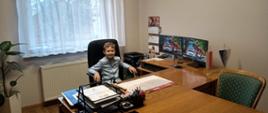 Za biurkiem, na fotelu siedzi ubrany w niebieską koszulę chłopiec