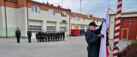 Strażacy stoją na baczność w dwóch rzędach oddając honor podczas wciągania flagi Polski na maszt przez poczet flagowy.