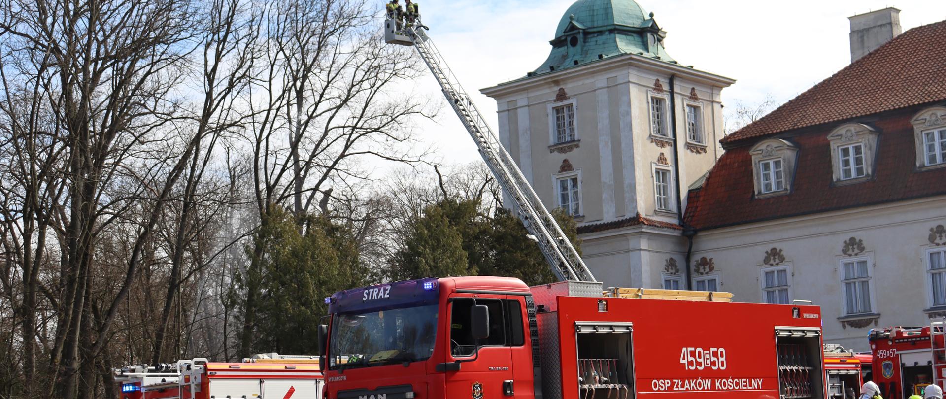 Na zdjęciu widzimy czerwone samochody strażackie na tle lasu i budynku muzeum. Możemy również dostrzec strażaków w koszy drabiny mechanicznej.