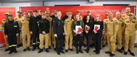 Grupa strażaków w umundurowaniu koloru piaskowego oraz komendant w ciemnym mundurze galowym stoją do wspólnego zdjęcia na tle czerwonej ścianki z napisem „Państwowa Straż Pożarna”. Na pierwszym planie trzy kobiety trzymają bukiety kwiatów i dyplomy.