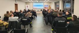 Widok z tyłu sali konferencyjnej podczas szkolenia – strażacy OSP siedzą na krzesłach i słuchają prelekcji prowadzonej przed ekranem z prezentacją.