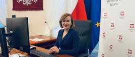 Sekretarz Stanu Anna Krupka siedząca przy biurku podczas wideokonferencji. W tle godło i flaga Polski, flaga UE oraz roll-up Ministerstwa Sportu.