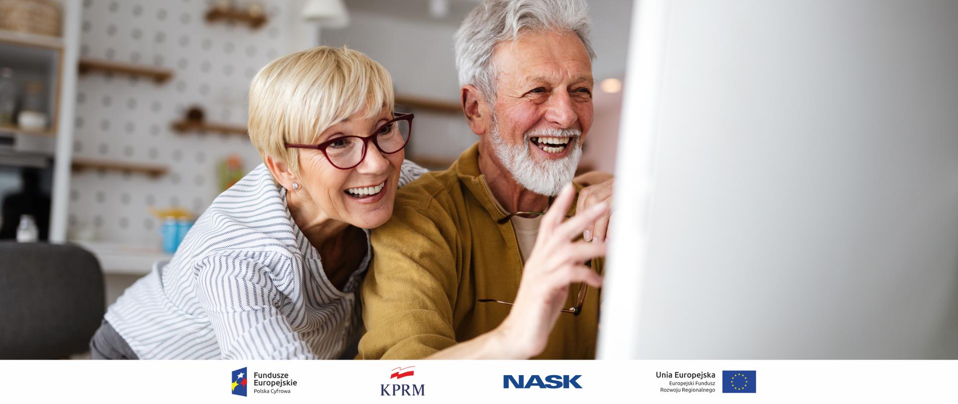 Dwoje szeroko uśmiechniętych seniorów przed komputerem.