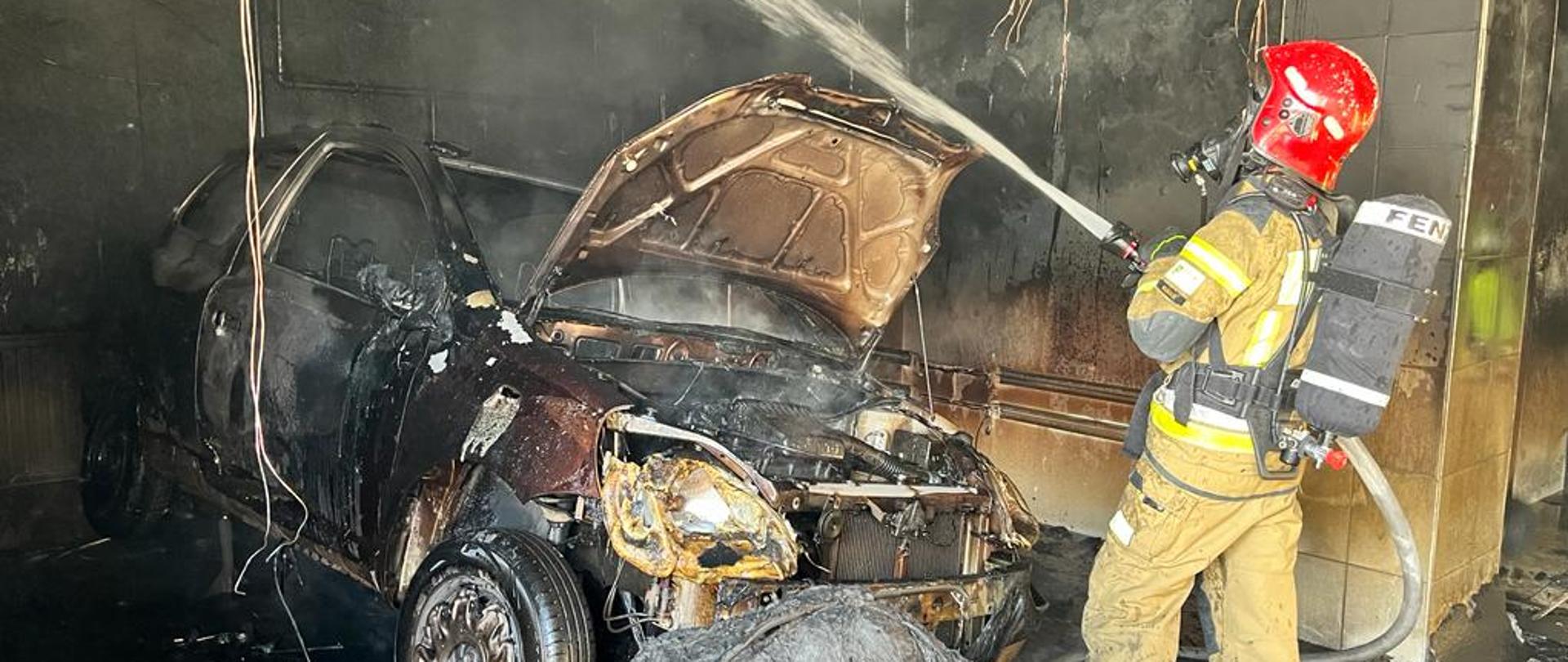 Po prawej strażak w aparacie ochrony układu oddechowego podający strumień wody na ścianę po lewej. Przed nim stoi spalony samochód, z podniesioną maską. Naokoło zwisają przewody instalacji elektrycznej.