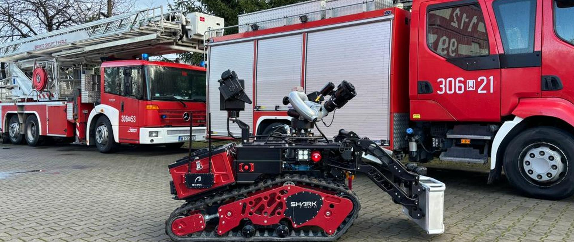 Robot gaśniczy Colossus na tle dwóch pojazdów pożarniczych: drabiny mechanicznej oraz średniego pojazdu gaśniczego