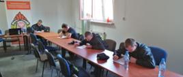 Na zdjęciu widać w sali wykładowej w KP PSP w Ostrowcu Św. 5 druhów OSP, którzy piszą egzamin pisemny. W tle godło RP i logo PSP. Druhowie ubrani w umundurowanie koszarowe.