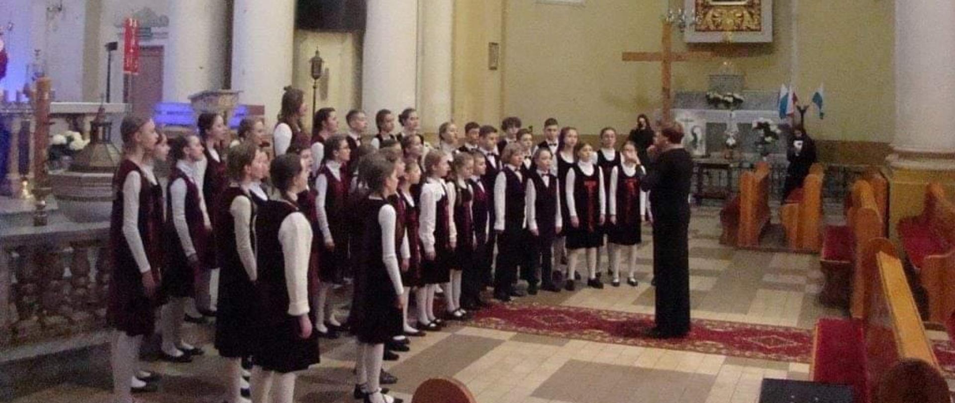 na zdjęciu w tle kościoła kilkunastoosobowy chór uczniowski drygowany przez nauczyciela
