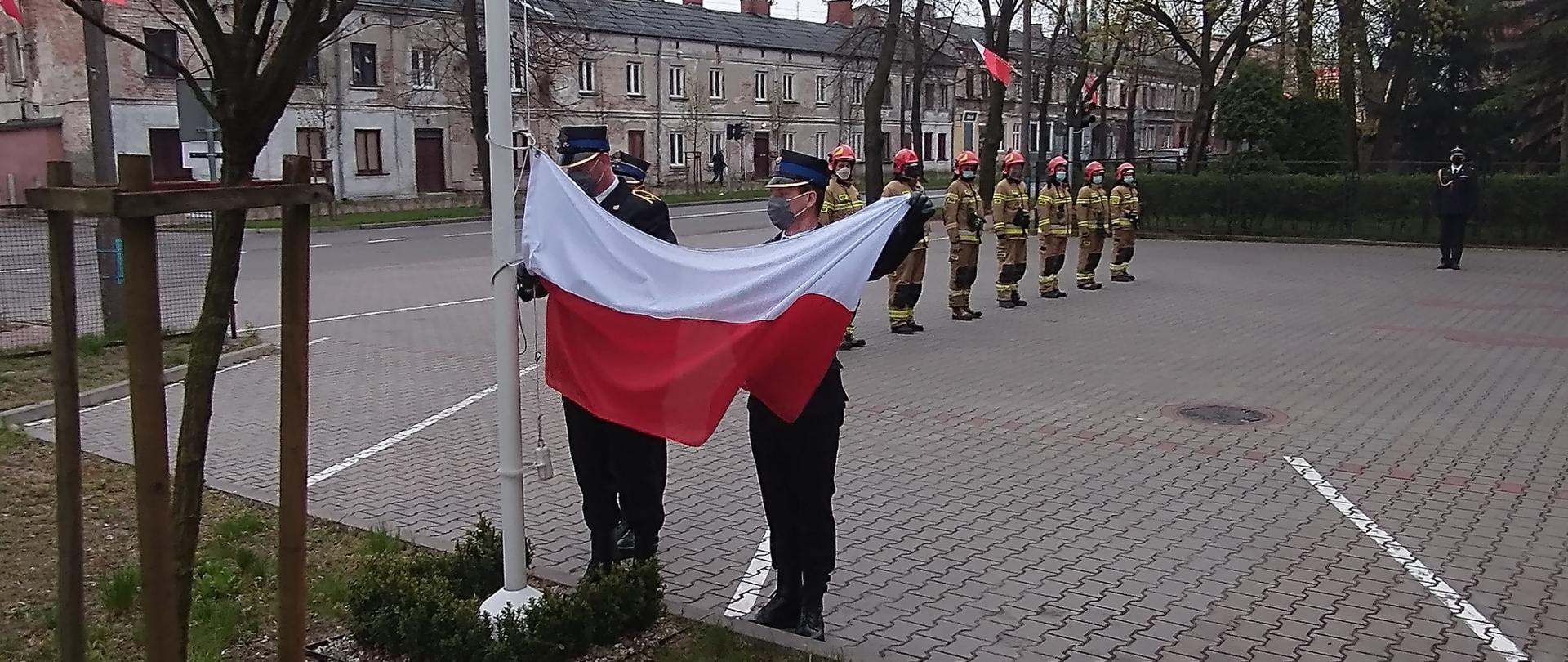 Poczet flagowy podnosi flagę na maszt. W tle stoją na baczność w szeregu strażacy w mundurach bojowych i komendant powiatowy PSP w Żyrardowie bryg. Tomasz Cybul