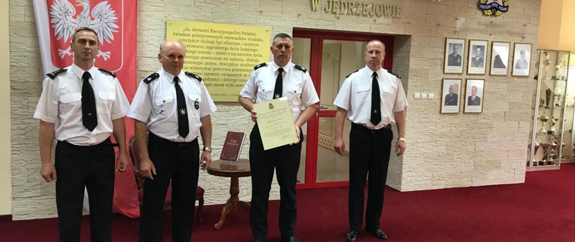 Zdjęcie przedstawia Komendanta Powiatowego PSP w Jędrzejowie- st. bryg. Pawła Jakubowskiego wraz ze swoim zastępcą- bryg. Tomaszem Szymańskim, którzy wręczają przedstawicielom OSP Brzegi decyzję o włączeniu do krajowego systemu ratowniczo-gaśniczego. Wszyscy strażacy w umundurowaniu galowym znajdują się na sali tradycji Komendy Powiatowej PSP w Jędrzejowie.