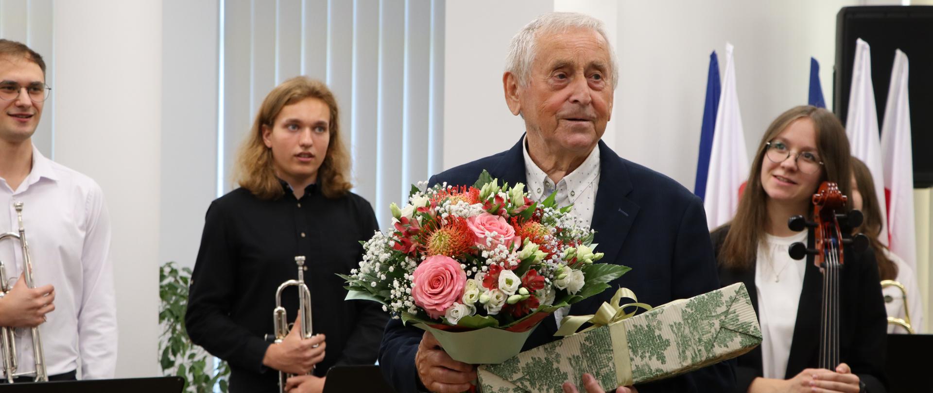 Mężczyzna ubrany w granatowy garnitur stoi na środku sali. W rękach trzyma bukiet kwiatów oraz pudełko z prezentem. Za nim stoi dziewczyna i dwóch chłopców. Chłopcy w rękach trzymają trąbki. Dziewczyna trzyma wiolonczele.