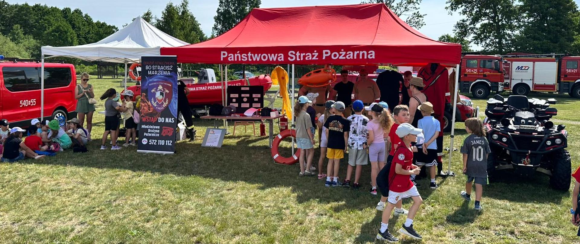 W tle dzieci, strażacy pokazujący sprzęt, widać pojazdy pożarnicze.