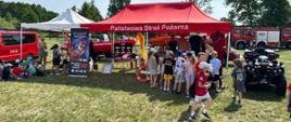 W tle dzieci, strażacy pokazujący sprzęt, widać pojazdy pożarnicze.