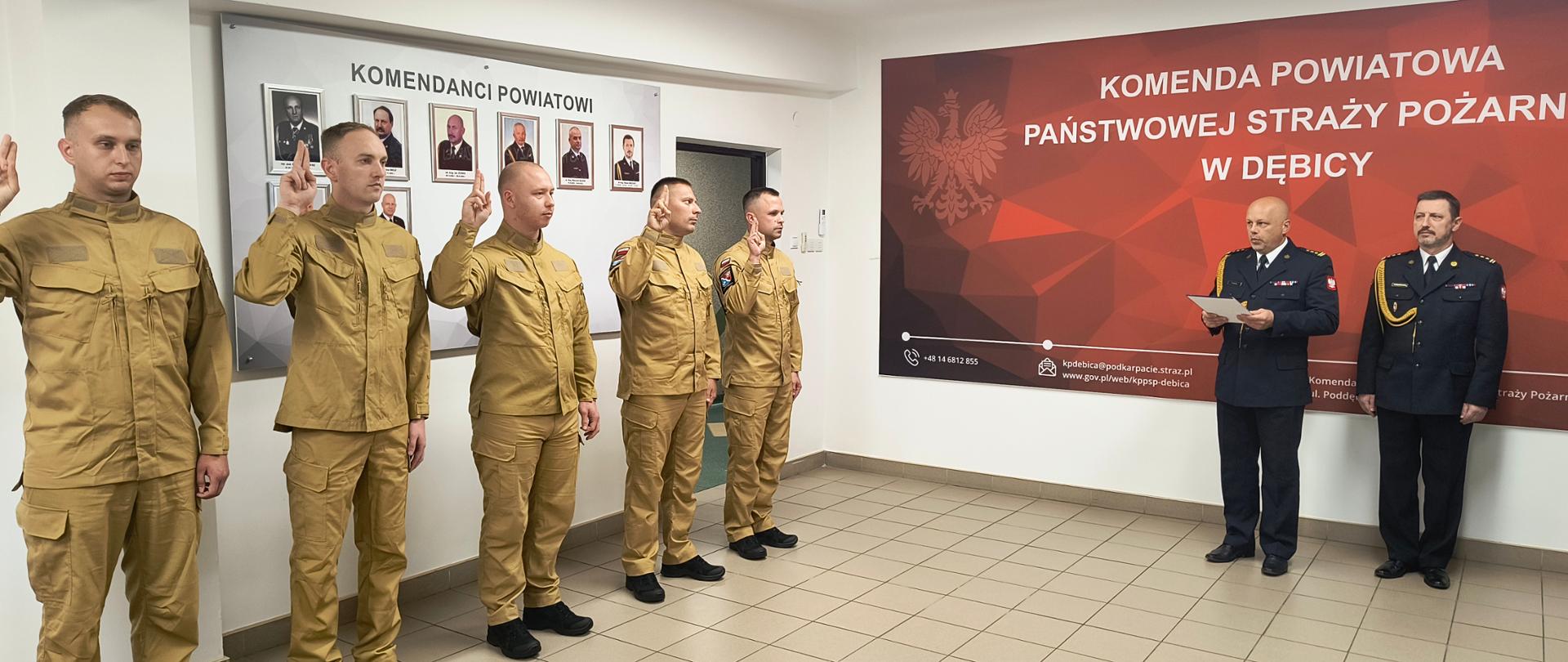 Strażacy ubrani w mundury wyjściowe stoją w sali konferencyjnej. Po lewej stronie, przy ścianie, stoi pięciu strażaków w jednym szeregu, którzy składają ślubowanie. Ci strażacy mają uniesione prawe ręce, przy czym dwa palce to jest wskazujący i środkowy są wyprostowane, a pozostałe palce mają zgięte. Po prawej stronie zdjęcia stoi komendant, który czyta rotę ślubowania. Obok komendanta stoi jego zastępca.