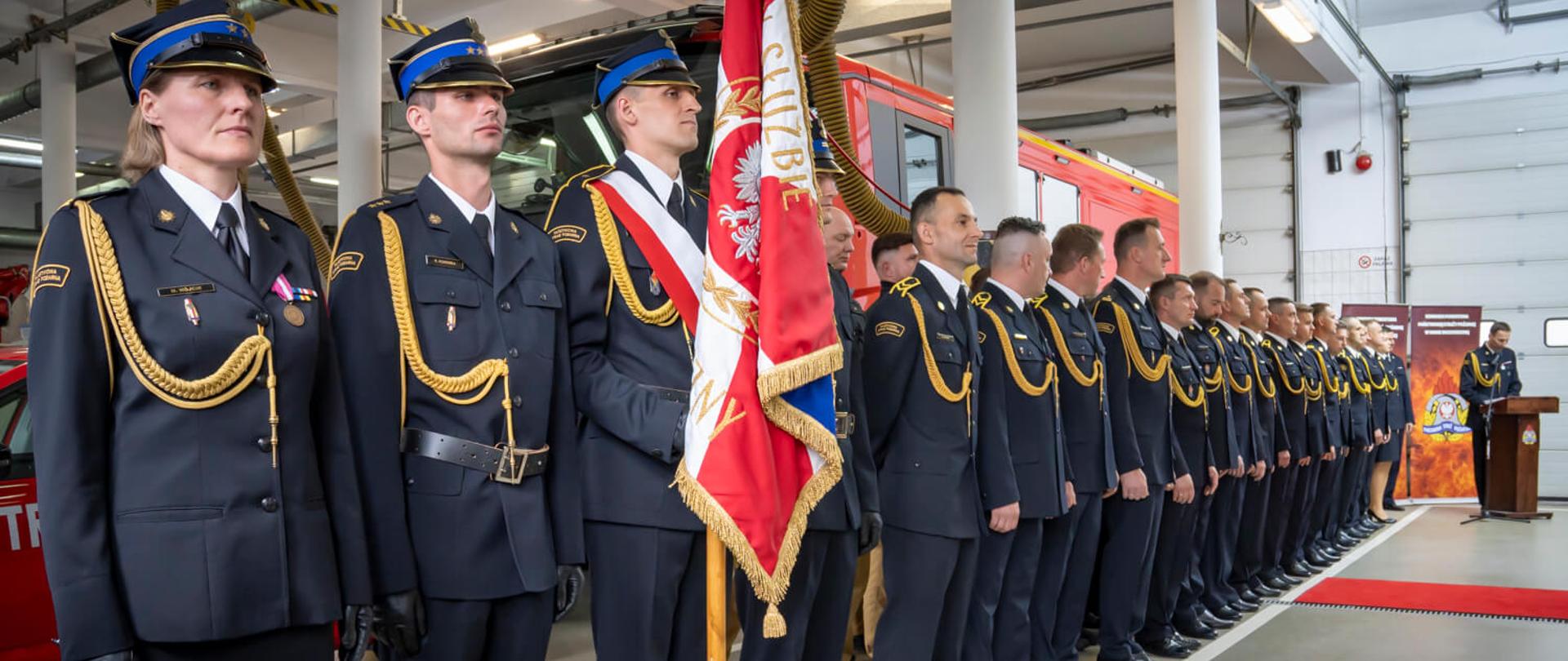 Formacja strażaków w galowych mundurach stoi na baczność podczas oficjalnej ceremonii w komendzie. Na pierwszym planie widoczny poczet sztandarowy z polskim sztandarem z napisem „Bogu na chwałę, ludziom na ratunek”. W tle znajduje się mównica oraz dekoracje tematyczne związane ze strażą pożarną.