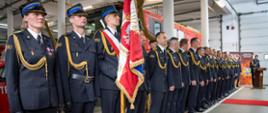 Formacja strażaków w galowych mundurach stoi na baczność podczas oficjalnej ceremonii w komendzie. Na pierwszym planie widoczny poczet sztandarowy z polskim sztandarem z napisem „Bogu na chwałę, ludziom na ratunek”. W tle znajduje się mównica oraz dekoracje tematyczne związane ze strażą pożarną.
