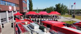 Na pierwszym planie widoczne dachy samochodów gaśniczych, dalej strażacy ustawieni w rzędach. Po lewej stronie przy bramie garażowej mały czerwony namiot, przy namiocie poczet flagowy. Na ostatnim planie zgromadzeni goście pod czerwonym namiotem. 