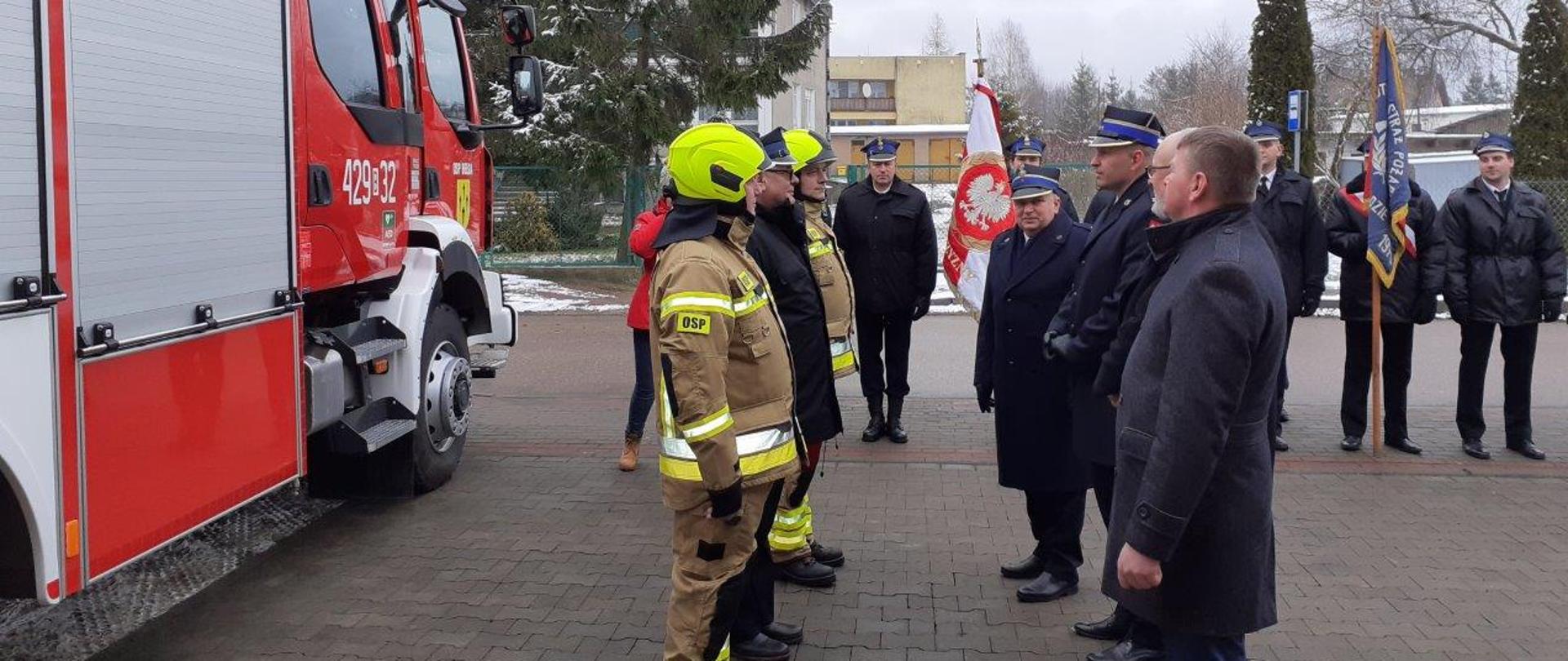 Moment przekazania pojazdu ratowniczego dla strażaków OSP Bełda ubranych w ubrania specjalne i hełmy.