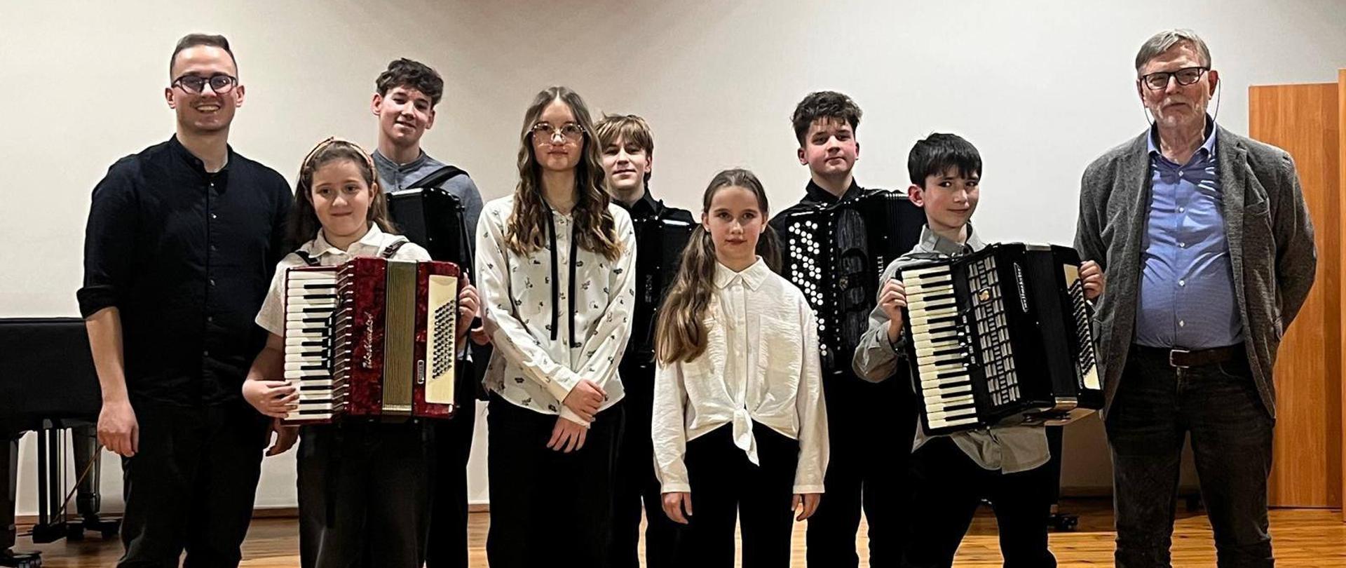 Pamiątkowe zdjęcie grupowe na scenie. Grupa siedmiu młodych muzyków (część z nich trzyma akordeony) stoi w rzędzie obok dwóch dorosłych mężczyzn – instruktorów. Wszyscy uśmiechają się w stronę aparatu. Sala ma jasne ściany i błyszczącą, drewnianą podłogę.