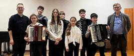 Pamiątkowe zdjęcie grupowe na scenie. Grupa siedmiu młodych muzyków (część z nich trzyma akordeony) stoi w rzędzie obok dwóch dorosłych mężczyzn – instruktorów. Wszyscy uśmiechają się w stronę aparatu. Sala ma jasne ściany i błyszczącą, drewnianą podłogę.