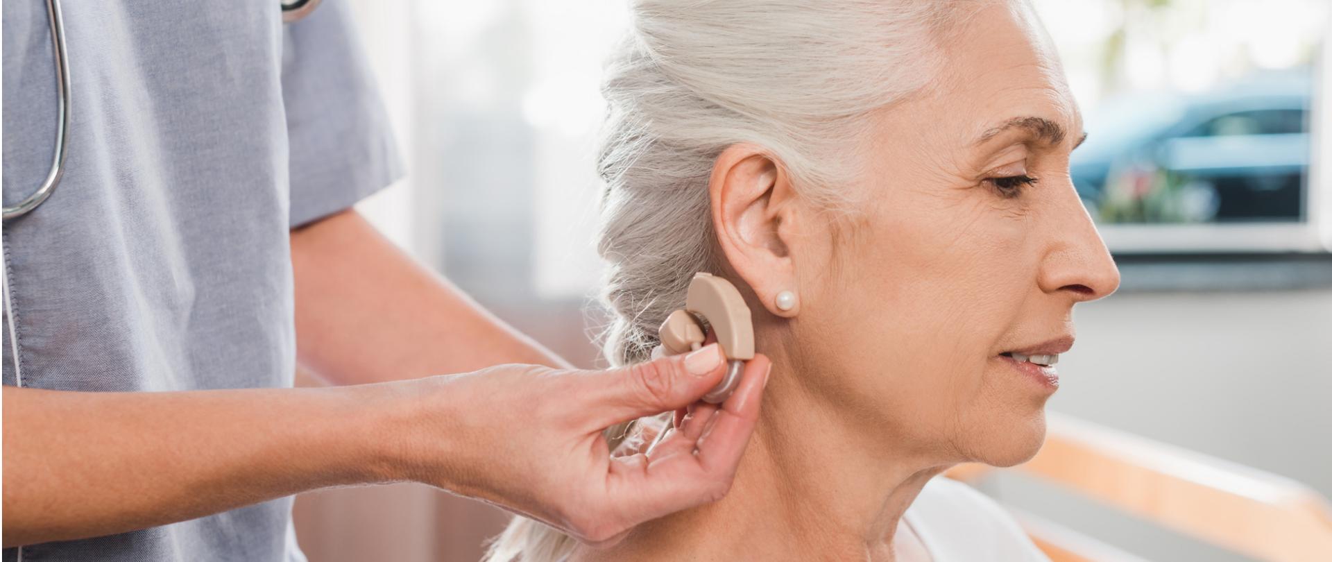 Na zdjęciu jest starsza kobieta i lekarka, która w dłoni trzyma aparat słuchowy.