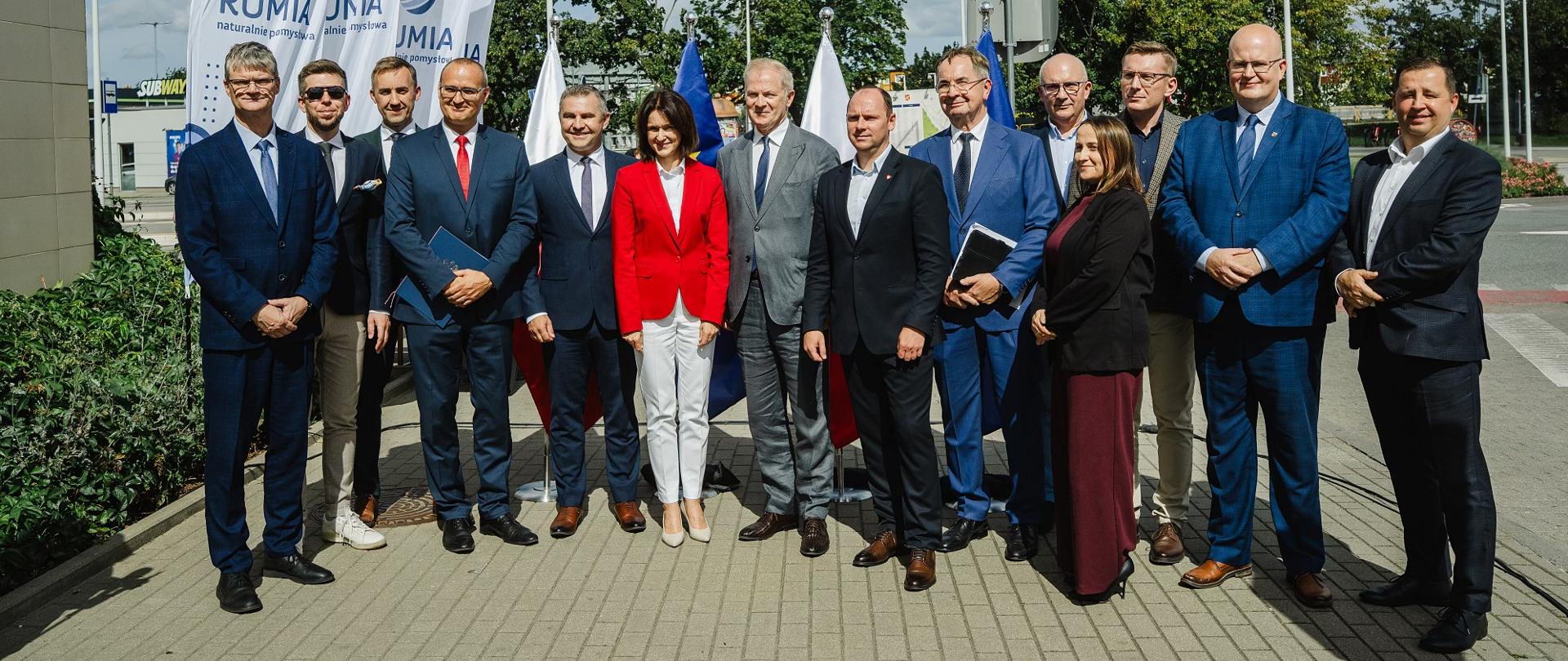 grupa osób stoi na placu, część trzyma teczki z dokumentami