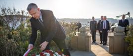 Na zdjęciu po lewej stronie minister Tomasz Siemoniak poprawiający biało-czerwoną szarfę na wieńcu. Wieniec w kolorach: czerwonym (róże), białym (gerbery) i zielonym. Po prawej stronie zdjęcia widać pięciu stojących mężczyzn (jeden w mundurze). W tle widać instalacje do wydobycia ropy naftowej (kiwony), samochody, roślinność oraz betonowe ogrodzenie miejsca pamięci.