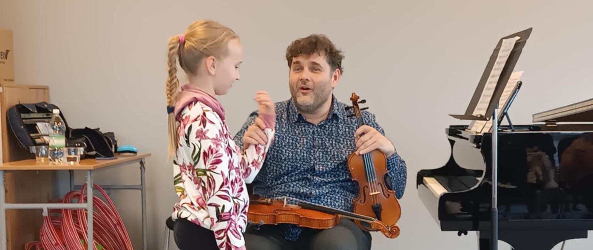 Na zdjęciu uchwycono ciepłą i zaangażowaną scenę z warsztatów skrzypcowych, pokazującą moment nauki między nauczycielem a młodą uczennicą. W centrum kadru siedzi prowadzący warsztaty na krześle, trzymając skrzypce na kolanach. Ma krótkie, kręcone włosy i ubrany jest w niebieską koszulę w drobny wzór oraz ciemne spodnie. Jego wyraz twarzy jest pogodny i pełen zaangażowania — wygląda, jakby właśnie coś tłumaczył lub odpowiadał na pytanie. Przed nim stoi młoda dziewczynka, uczennica. Ma blond włosy związane w długi warkocz i nosi kolorową bluzę z motywem liści oraz ciemne legginsy z napisem wzdłuż nogawki. Dziewczynka uważnie patrzy na nauczyciela i wydaje się słuchać go z zaciekawieniem, być może trzyma jego dłoń w ramach ćwiczenia lub instrukcji dotyczącej uchwytu smyczka. W tle znajduje się czarny fortepian z otwartą pokrywą i pulpit z nutami, co sugeruje, że zajęcia odbywają się w sali muzycznej. Obok fortepianu widać stolik z wodą i materiałami, a w kącie pomieszczenia – kartony i czerwone obręcze, być może elementy wykorzystywane w pracy z dziećmi. Scena emanuje ciepłem, cierpliwością i skupieniem, charakterystycznym dla zajęć indywidualnych, gdzie nauczyciel przekazuje uczniowi wiedzę w bezpośrednim, wspierającym kontakcie.