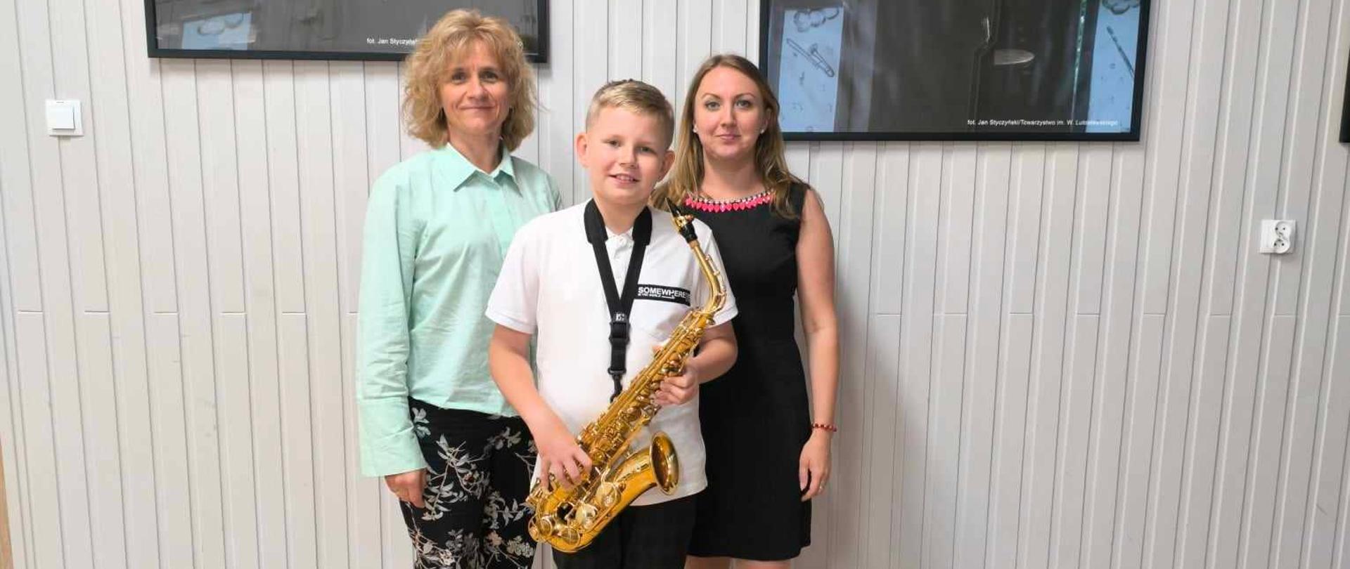Uczeń trzymający saksofon oraz nauczycielka saksofonu i akompaniatorka stoją w auli PSM. W tle widnieją dwie czaro-białe fotografie Witolda Lutosławskiego. Ogólna kolorystyka – czarno-biała.