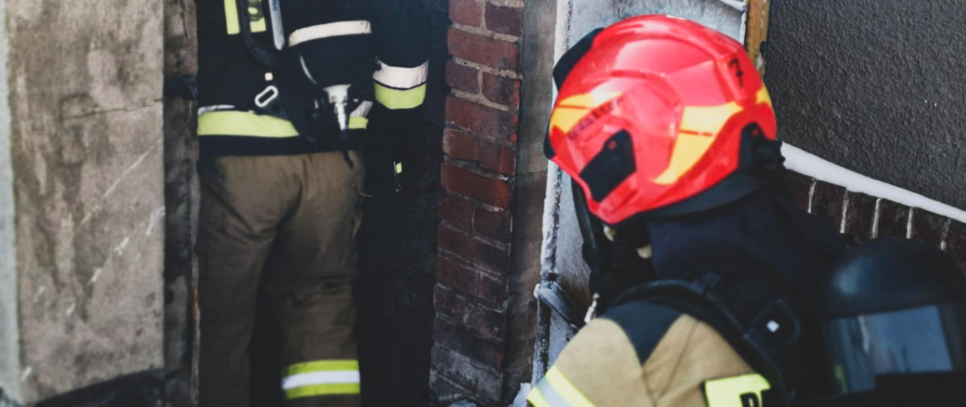 Na zdjęciu znajduje się dwóch strażaków ubranych w aparaty powietrzne i ubrania specjalne, którzy schodzą po schodach do piwnicy w budynku mieszkalnym
