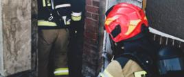 Na zdjęciu znajduje się dwóch strażaków ubranych w aparaty powietrzne i ubrania specjalne, którzy schodzą po schodach do piwnicy w budynku mieszkalnym
