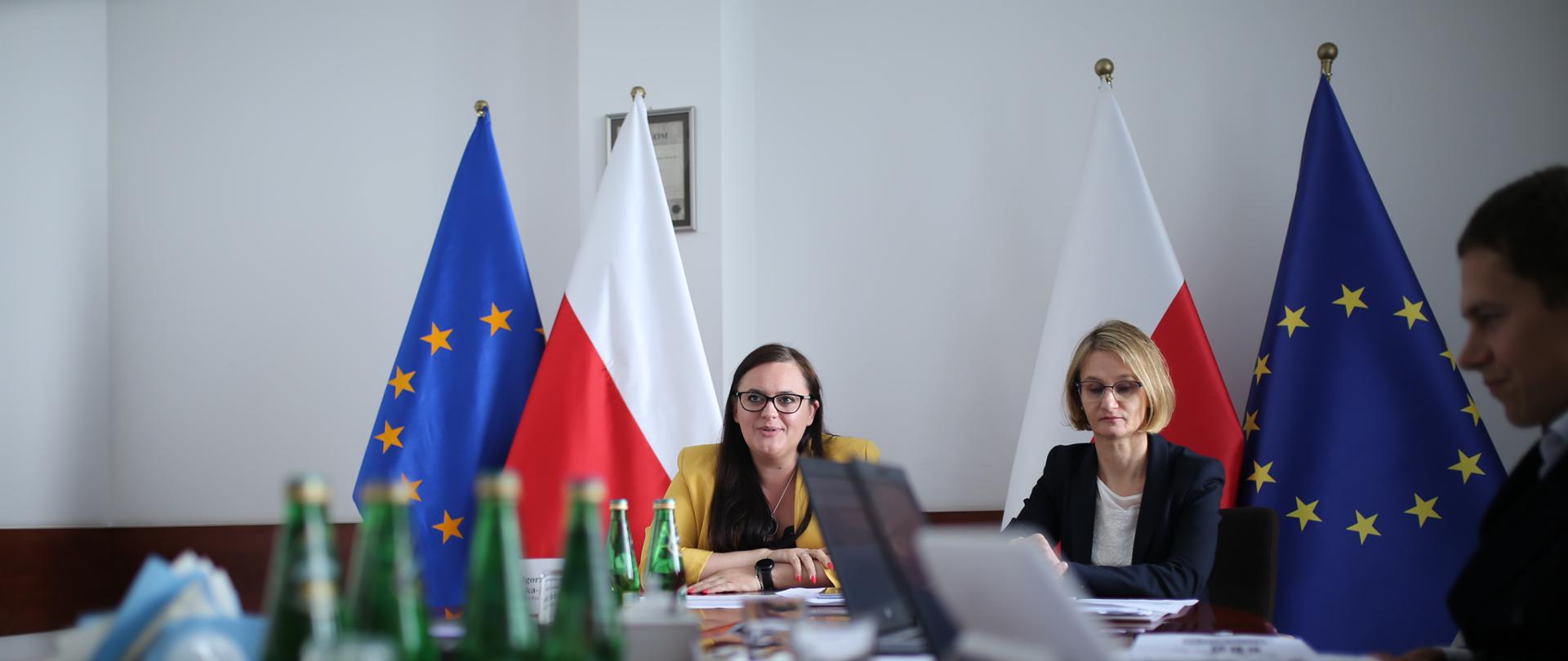 Na zdjęciu trzy osoby siedzą przy stole konferencyjnym, za nimi flagi Polski i Unii Europejskiej