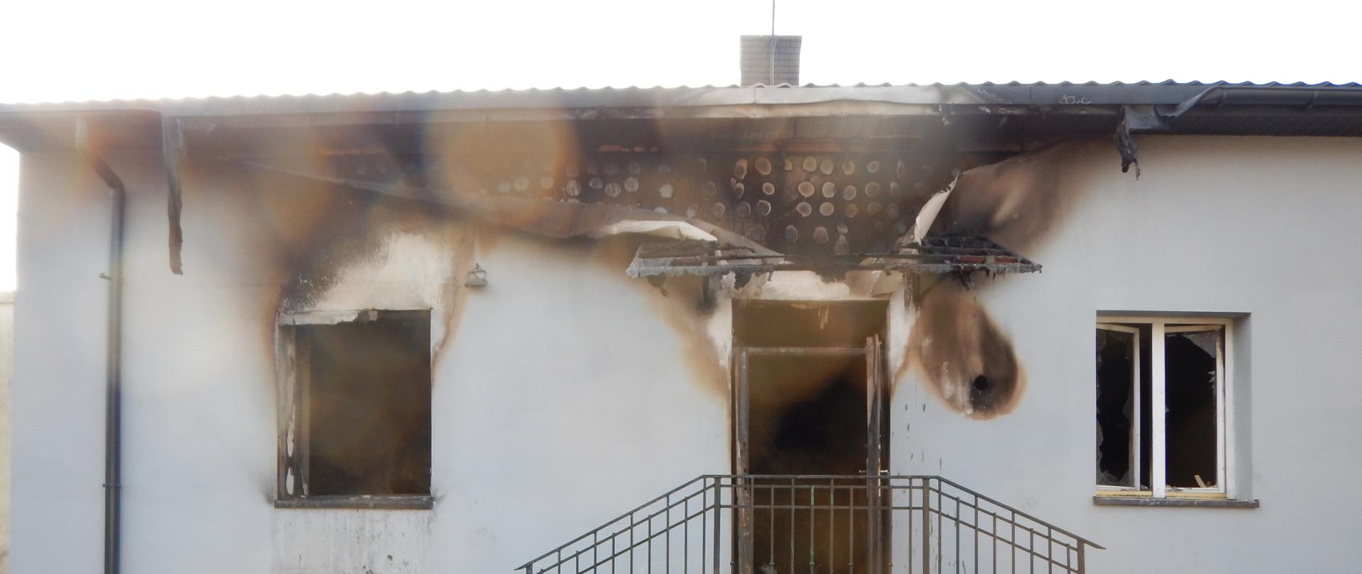 Zdjęcie przedstawia budynek mieszkalny, który w wyniku pożaru ma całkowicie zniszczone dwa pomieszczenia na parterze oraz część poddasza.