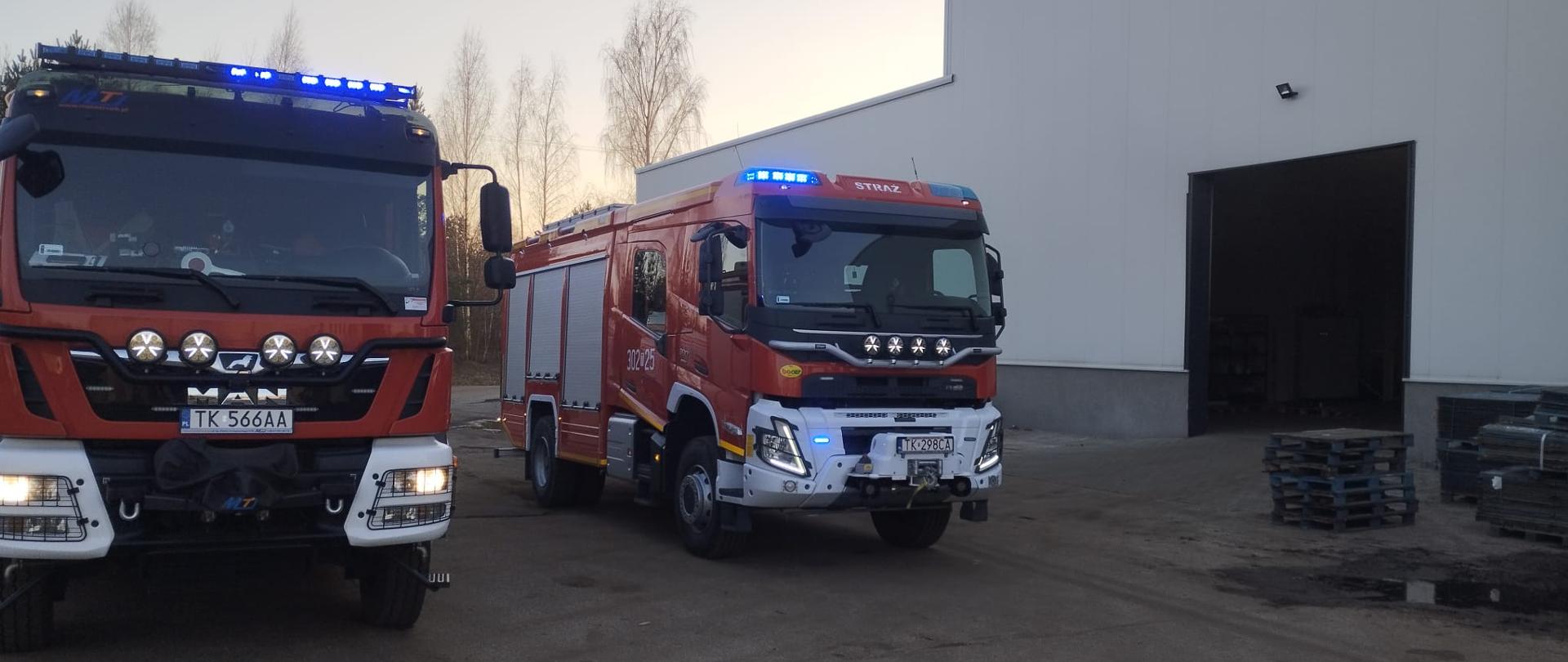 Zdjęcie przedstawia dwa samochody pożarnicze stojące na placu przy hali produkcyjnej. Po prawej stronie: hala przemysłowa gdzie doszło do pożaru.