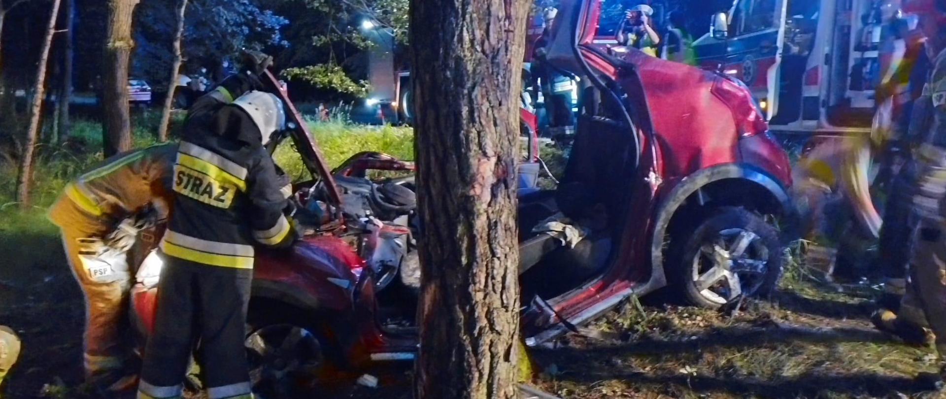 przy rozbitym samochodzie który ma wyięty i odgięty dach stoją dwaj strażacy zdjęcie wykonane nocą miejsce zdarzenia jest oświetlone
