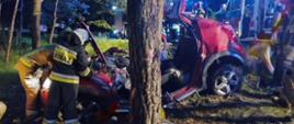 przy rozbitym samochodzie który ma wycięty i odgięty dach oraz wycięte drzwi stoją dwaj strażacy zdjęcie wykonane nocą miejsce zdarzenia jest oświetlone