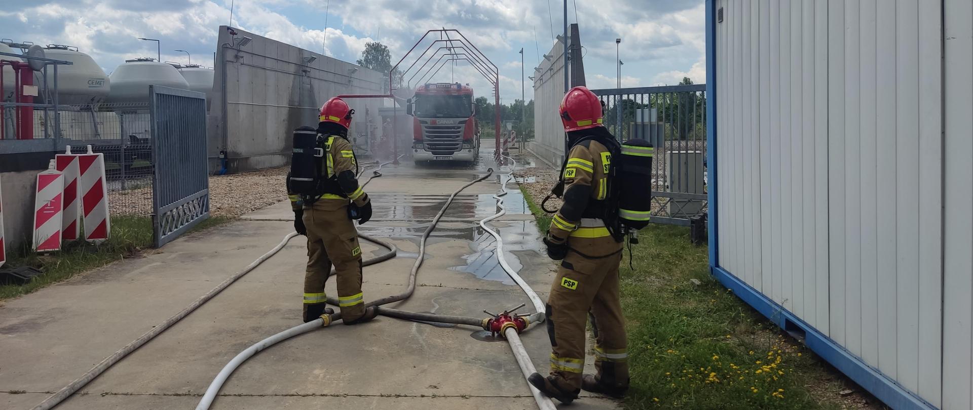 Na zdjęciu dwóch strażaków w umundurowaniu specjalnym oraz sprzęcie ochrony układu oddechowego. W tle autocysterna na stanowisku do rozładunku autocystern, obok niej rozstawiona kurtyny wodne. Pod nogami strażaków rozwinięte węże doprowadzające wodę do kurtyn. 