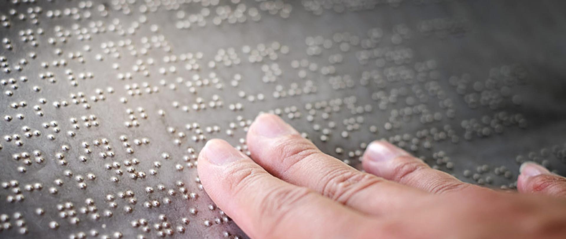 Dłoń odczytująca tekst zapisany alfabetem Braille’a na wypukłej tablicy.