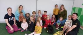 Na zdjęciu znajdują się dzieci siedzące na podłodze, trzy opiekunki oraz strażak