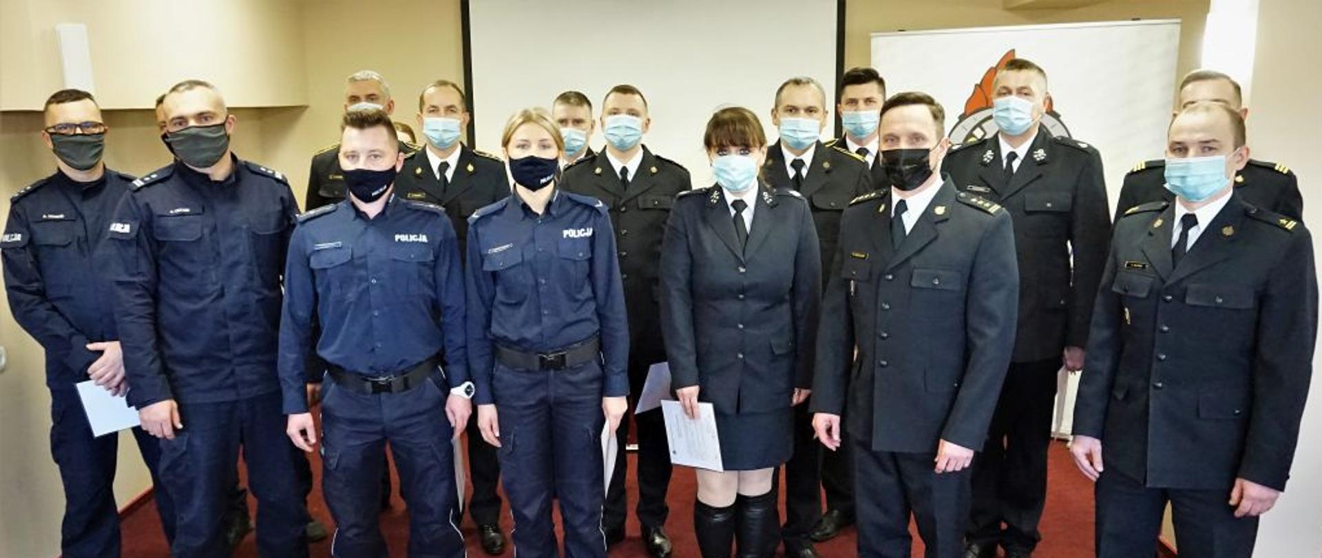 W sali na czerwonej wykładzinie w dwóch rzędach stoją funkcjonariusze policji straży pożarnej oraz druhowie OSP w granatowych mundurach w maseczkach. 