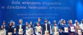 W świetle niebieskich reflektorów grupa młodzieży ubrana na galowo trzyma w rękach teczki z dyplomami. W tle napis: "Gala wręczenia stypendiów w dziedzinie twórczości artystycznej uczniom uzdolnionym artystycznie z województwa podlaskiego" 