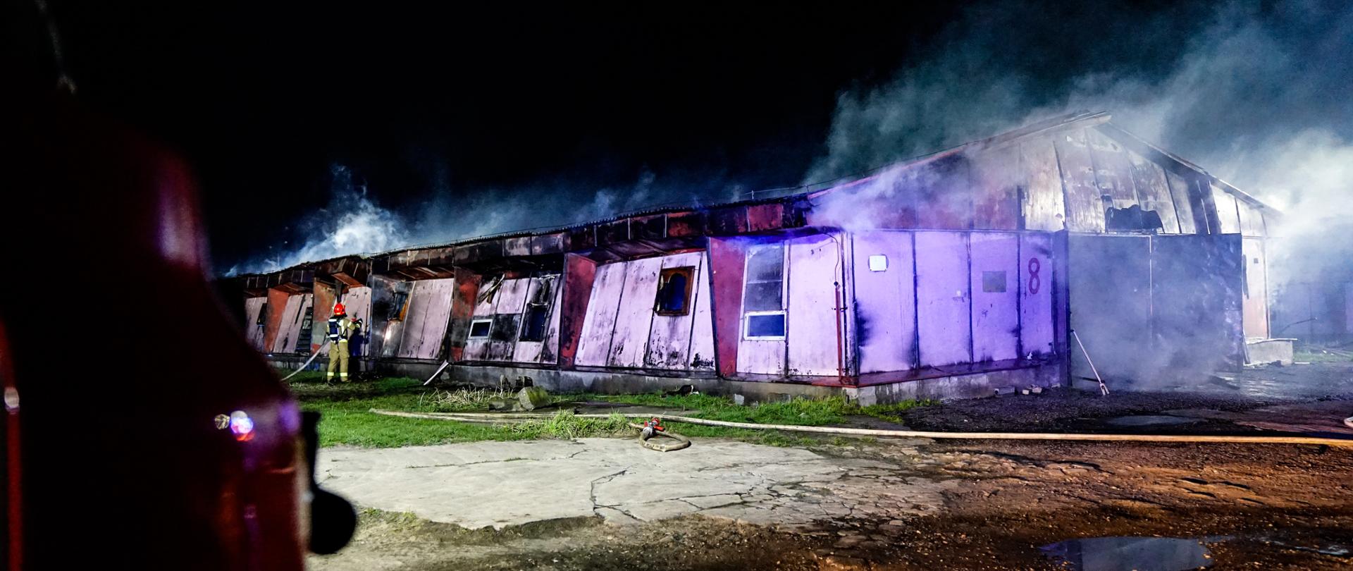 Na zdjęciu widać budynek w którym prowadzone są działania gaśnicze, spod dachu wydobywający się dym, obok budynku strażak PSP w ubraniu specjalnym prowadzący działania gaśnicze 