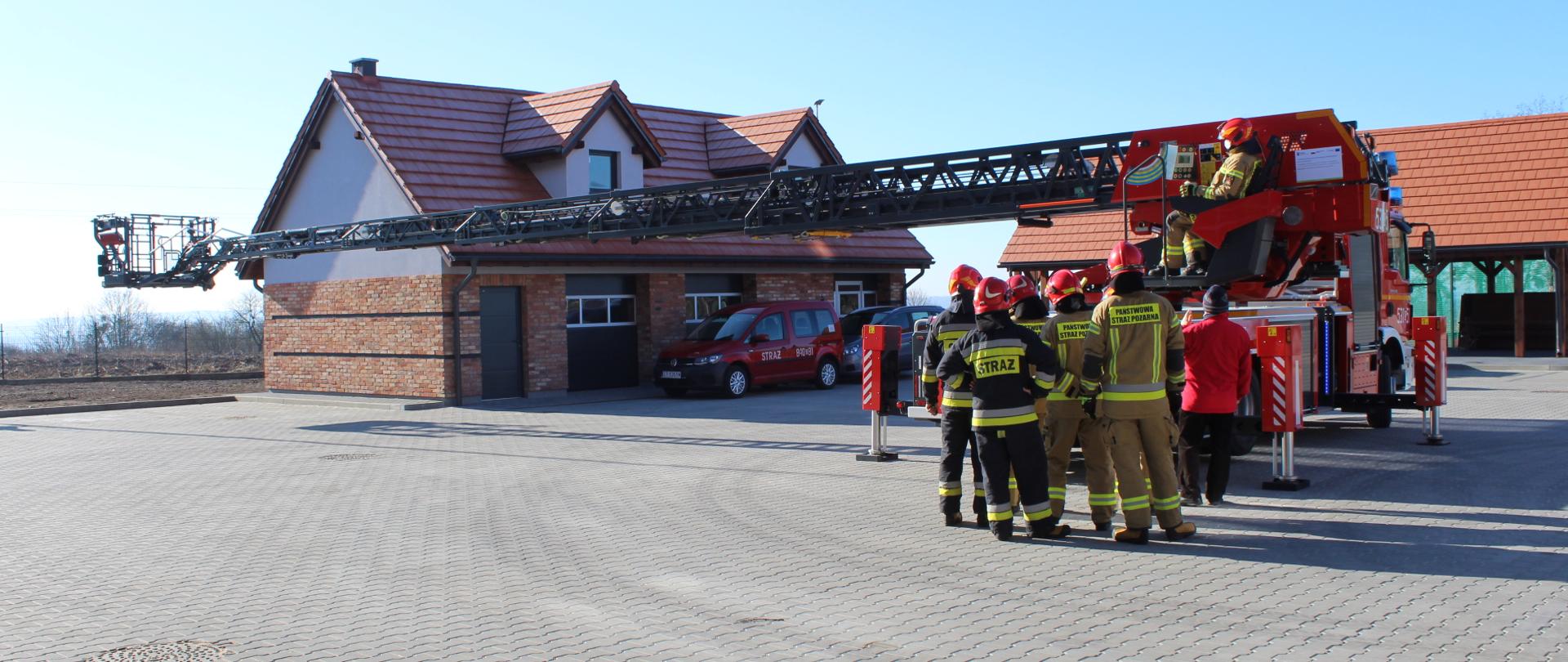 Na placu przed budynkiem Komendy Powiatowej Państwowej Straży Pożarnej w Świeciu stoi samochód z drabiną mechaniczną tyłem, z rozłożonym w poziomie zespołem drabin na odległość około 18 metrów i przesuniętym o 90 stopni w lewo. Przed nim odwróceni tyłem grupa strażaków ubranych w ubrania specjalne i hełmy oraz egzaminator.