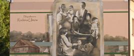 Widok na ścianę żółtego budynku, na której namalowany jest mural - odwzorowanie czarni-białego zdjęcia rodziny z sześciorgiem dzieci i napis Błogosławiona rodzina Ulmów.