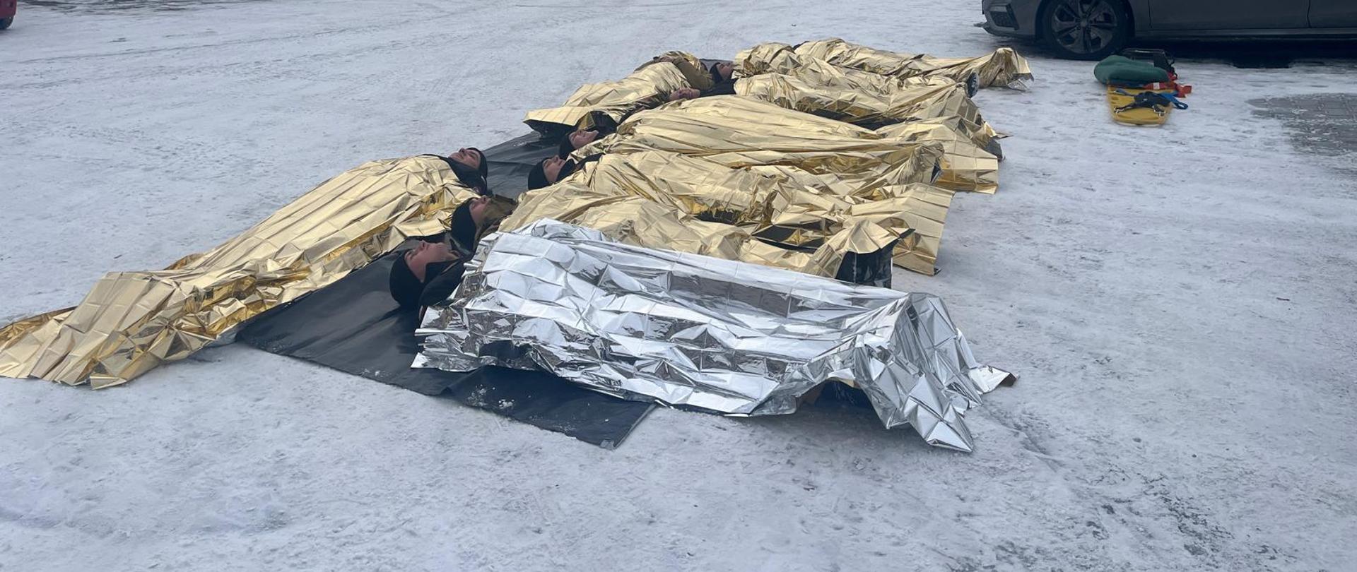 Teren pomiędzy budynkami CS PSP, grupa kadetów okrytych folią życia leży na macie rozłożonej na betonie pokrytym śniegiem