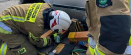 Dwóch strażaków Ochotniczej Straży Pożarnej w umundurowaniu specjalnym wykonuje stabilizację uszkodzonego pojazdu za pomocą drewnianych klinów oraz sprzętu pneumatycznego. W tle zaparkowane wraki aut i budynek mieszkalny. Niebo zachmurzone.
