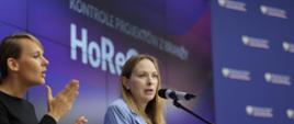 In the photo, Minister Katarzyna Pełczyńska-Nałęcz stands on stage in front of a microphone. A sign language interpreter stands next to her